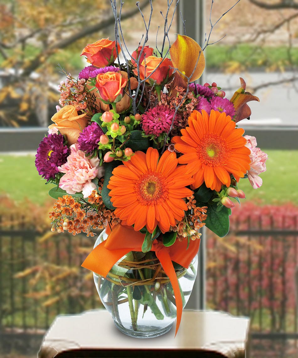 Gerbera daisies, asters, calla lilies and other flowers in a clear glass vase tied with an orange bow.